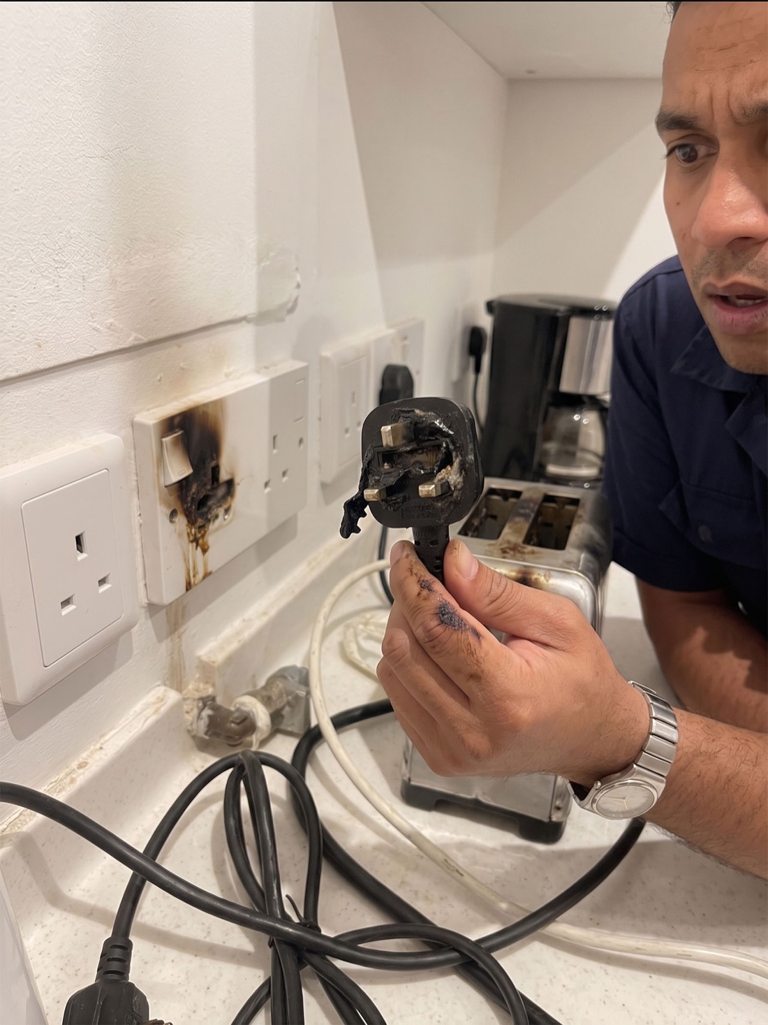 A burnt, melted three-pin plug held next to a scorched, charred wall socket
